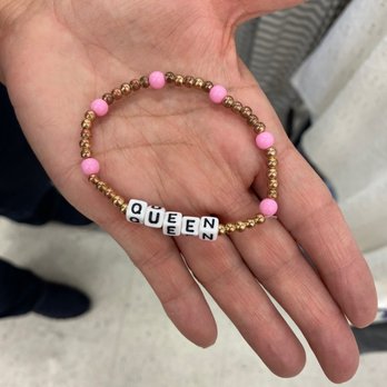 JULIA - A beaded bracelet featuring pink, gold, and white beads spelling out "QUEEN" is displayed in a person's hand. The bracelet is composed of small round beads, with larger pink beads interspersed among gold beads, and white letter beads forming the word. The person holding the bracelet is wearing a watch on their wrist. The background appears to be an indoor setting, possibly a store or public space, with blurred elements and a tiled floor. - Jewelry Near Me - Fresno, California