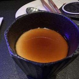 Chawanmushi - A black ceramic cup containing a brown liquid, possibly tea or broth, placed on a dark surface with utensils and a napkin nearby.