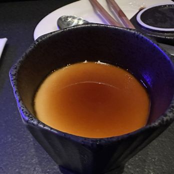 Nohri - Chawanmushi - A black ceramic cup containing a brown liquid, possibly tea or broth, placed on a dark surface with utensils and a napkin nearby. - Sushi Bars Near Me - Boston, Massachusetts