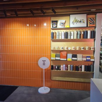 Window Coffee Bar - The image shows a retail store interior with a wooden shelf unit mounted on an orange tiled wall. The shelves hold various mugs, bottles, and framed artwork. A sign on a stand reads 'COME AS YOU ARE.' The ceiling has exposed wooden beams and track lighting. The floor is concrete, and part of a white counter is visible on the right. - Coffee & Tea Near Me - Phoenix, Arizona