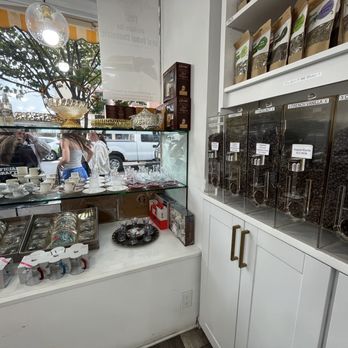 Apricot Nuts - The image shows an interior of a shop selling coffee beans and tea-related products. On the right, there are four dispensers filled with different types of coffee beans, labeled with their varieties and prices. Below the dispensers, there are white cabinets. To the left, a glass display case contains tea cups, mugs, and other small items. The shop has a large window at the front, through which parked cars and people walking outside are visible. The shop interior includes shelves with packaged products and decorative items. - Desserts Near Me - Westfield, New Jersey