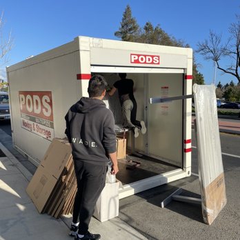 A2B Movers - Two people are engaged in loading or unloading a PODS moving and storage container situated on a sidewalk. One person stands nearby holding boxes, while the other is inside the container, arranging or retrieving items. The container is partially open, with some cardboard boxes and a large panel leaning against it. The scene takes place outdoors on a clear day, with trees and parked cars visible in the background. - Movers Near Me - San Jose, California