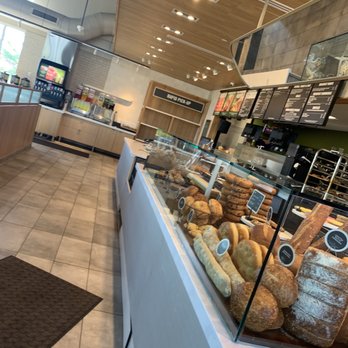 Panera Bread - Bakery... - Inside a cafe, a bakery display case showcases different types of bread and baked items, with a menu board overhead and a beverage station in the background. - Sandwiches Near Me - Wheaton, Illinois