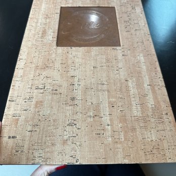 Evie’s - Menu - A rectangular cork-textured menu with a metallic square logo in the center, held by a person with red nail polish, on a dark table. - Beer, Wine & Spirits Near Me - Westerly, Rhode Island