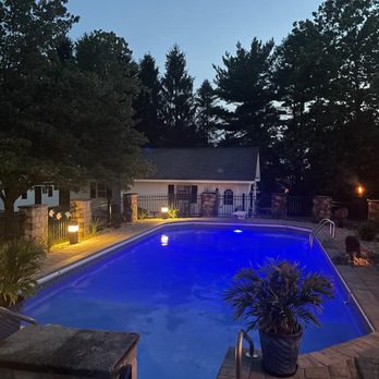 Solid Brick Contracting - A backyard swimming pool is illuminated with blue underwater lights during dusk, with surrounding potted plants, outdoor lighting, and trees in the background. The pool area is enclosed by a fence and features a paved deck with steps leading into the water. - Landscape Architects or Designers Near Me - Aberdeen, Maryland