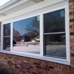 2 new double hung windows with a fixed pane in the middle. - The image shows a large, double-hung window with a white frame installed on a brick house exterior. The window has two sections, with the left side slightly open. The reflection in the glass shows a snowy yard, trees, and neighboring houses. A wall-mounted outdoor light fixture is visible on the right side of the window, along with a sign that reads 'No Soliciting.' The scene suggests a residential property during winter.