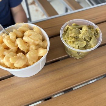 City Barbeque - A white disposable cup contains macaroni and cheese with a spoon inside, and a clear plastic cup holds chopped pickles. Both containers are on a wooden table with a blurred background showing part of a person and some outdoor seating. - Barbeque Near Me - Lafayette, Indiana