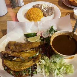 Quesabirria y Consomm - A plate featuring three tacos with grilled meat and vegetables, accompanied by shredded lettuce, radish slices, lime, and a bowl of dark sauce. In the background, a plate of rice with meat and beans is visible.