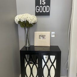 A black cabinet with patterned glass doors holds a vase of white hydrangeas and a framed photo, set against a gray wall with wall art.