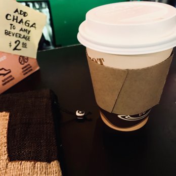 OK Cafe - Hot! (Almond Mylk Lavender London Fog Latte) Delicious - A disposable coffee cup with a plastic lid and a cardboard sleeve is positioned on a dark table. The background includes a green wall and a small sign offering add-ons for beverages. The scene suggests a cafe environment with items like a woven bag and a note about beverage add-ons visible nearby. - Coffee & Tea Near Me - Astoria, New York