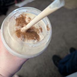 Snicker doodle malt - A person holds a clear plastic cup containing a milkshake topped with cinnamon powder. The milkshake appears creamy and light-colored, with a straw inserted. The background shows an indoor environment with a carpeted floor and some furniture or objects, but they are out of focus.