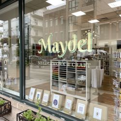 Maydel - The outside of a shop in a brick building with flower boxes outside and a painted gold window sign that says Maydel