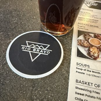 Rio Bravo Brewing - Squirrel nuts beer - A round Rio Bravo coaster, a tall glass of dark beer, and a menu are placed on a dark, textured table surface. - American Near Me - Albuquerque, New Mexico
