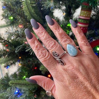 Gorgeous Nails - A close-up of a hand with dark gray matte nail polish and multiple rings, including a large teardrop-shaped blue stone ring and a silver ring with a spiral design, is shown in front of a Christmas tree decorated with multicolored lights and ornaments. The background features a Christmas tree with green branches, colorful lights, and a wrapped gift ornament. - Nail Salons Near Me - Frederick, Maryland