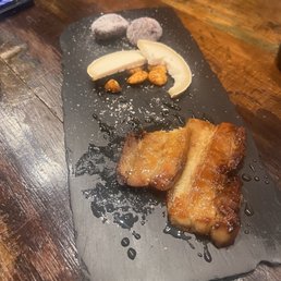 Pork Belly - A dessert presentation on a black slate includes two fried banana pieces, a slice of cheese, candied almonds, and two purple mochi-like sweets dusted with powdered sugar. The items are arranged with some drizzled syrup, and the setting appears to be in a restaurant with a wooden table.