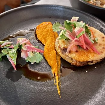 Root & Flower - The image shows a plated dish with a grilled patty topped with herbs and vegetables, including cilantro and pink radish slices. A dollop of orange sauce is placed beside the patty, with a drizzle of dark sauce on the plate. The dish is presented on a black plate, highlighting the colorful ingredients and garnishes. - Wine Bars Near Me - Vail, Colorado