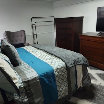 Ultimate Treasures - A bedroom features a bed with patterned bedding, a dresser, a TV on top, and a gray upholstered chair, with a plain white wall and a rug. - Antiques Near Me - Downers Grove, Illinois