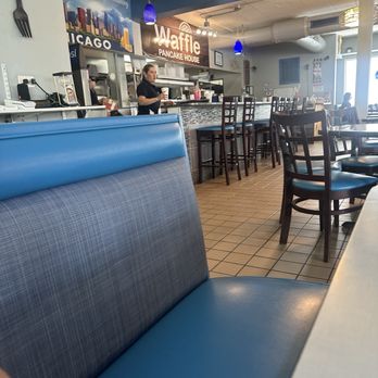Waffle Pancake House - Server + kitchen/bar area. - The image shows the interior of a breakfast restaurant with a counter area where a staff member is preparing food. There are multiple chairs and booths for seating, with a blue booth in the foreground. The decor includes hanging lights, a sign for Waffle Pancake House, and a tiled floor. The restaurant appears to be well-lit with natural and artificial lighting. - Breakfast & Brunch Near Me - Chicago, Illinois