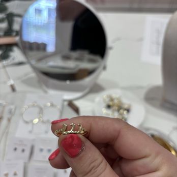 Anna Nails Studio - A person with red nail polish holds a gold ring featuring a crown design, displayed in a jewelry store with a mirror and jewelry on the table. - Nail Technicians Near Me - Villa Park, Illinois