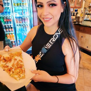 La Piazza - A woman with long dark hair, makeup, and a black sleeveless top is seated in a restaurant, holding a slice of pizza on a paper plate. Behind her are a refrigerator with bottled drinks and a wooden counter with a display case. She is smiling at the camera. - Pizza Near Me - Plainview, New York