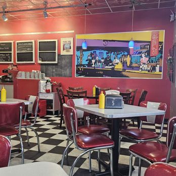 Graces Place - The round table - The image shows an interior of a retro 50's-themed diner with red walls and checkered black and white flooring. There are several red and white chairs around tables, with some tables having yellow condiment bottles. A large mural depicting a bar scene with people is on the wall, illuminated by blue pendant lights. The ceiling has a metallic patterned surface with spotlights and track lighting. A small counter area with a chalkboard menu and a soda fountain is visible in the background. - Diners Near Me - Cascade, Iowa