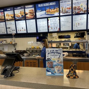 Culver's - The image shows the interior of a fast-food restaurant with a counter and a menu display overhead. The menu boards list various food items such as sandwiches, salads, seafood, chicken, and desserts. Decorations include plush toys and a promotional sign on the counter advertising ice cream flavors. Staff members are visible behind the counter, preparing orders. The setting includes a beige countertop, a cash register, and a tiled wall behind the counter. - Burgers Near Me - Springfield, Illinois