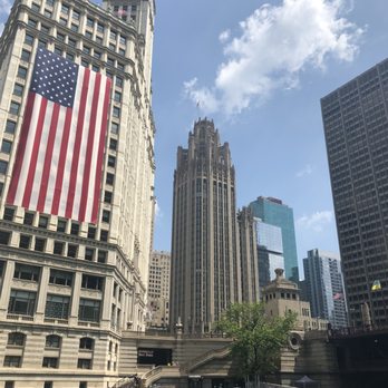 Chicago Architecture Center - Wrigley Building. - The image shows a cityscape of Chicago with prominent buildings including the Wrigley Building with a clock tower and a large American flag hanging on its facade. Other skyscrapers are visible in the background, and the scene is set along the Chicago River with water in the foreground. The sky is partly cloudy with blue sky and white clouds. - Historical Tours Near Me - Chicago, Illinois