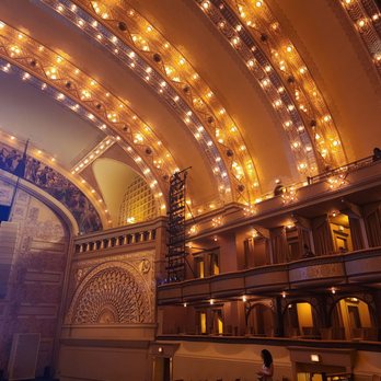 Auditorium Theatre - The image shows the interior of a historic theatre with elaborate architectural details. Gold-colored arches with numerous lights span across the ceiling, and there are balconies with seating areas. Murals and decorative patterns adorn the walls, emphasizing the theatre's ornate design. Audience members are seated in the lower section, and a few individuals are visible near the aisles and balconies, indicating a performance setting. - Performing Arts Near Me - Chicago, Illinois