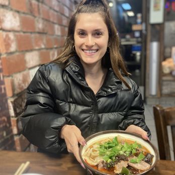 Wild Ginger - A woman with long brown hair in a high ponytail, wearing a black puffer jacket, sits at a wooden table in a restaurant, holding a bowl of spicy noodle soup garnished with herbs and meat, and smiles. - Chinese Near Me - Alameda, California