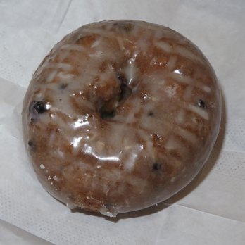 Kim’s Donuts - Blueberry - A round donut with a hole in the center, covered in a shiny, translucent glaze. The donut contains visible blueberry pieces embedded within, and it is placed on white paper. The surface appears moist and sugary, typical of a freshly glazed donut. - Donuts Near Me - Lowell, Massachusetts