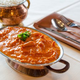 Butter Chicken - A serving of butter chicken with a rich, orange sauce is presented in a metal bowl, garnished with a small sprig of herbs. In the background, a copper pot filled with white rice is visible. The table setting includes a folded napkin with a fork, knife, and spoon placed on top, on a light-colored tablecloth.