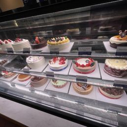 A glass display case contains multiple decorated cakes arranged on white trays, featuring fruit toppings, chocolate layers, and various frosting styles. The cakes are labeled with small signs indicating their flavors, such as strawberry, chocolate, and red velvet. The display is set in a bakery or cafe environment, with a countertop visible below the case.