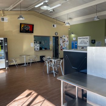 The Laundry Room - Modern decor and quaint. - Laundromat interior featuring seating areas, a soap center, a vending machine, and a wall-mounted TV, with natural light and ceiling lights. - Laundry Services Near Me - Moriarty, New Mexico
