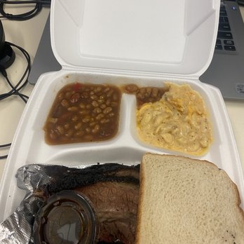 Texas Steele BBQ - A foam takeout box contains baked beans, macaroni and cheese, a slice of bread, and a cooked meat piece, placed on a desk with a laptop and cables. - Food Trucks Near Me - Columbus, Ohio
