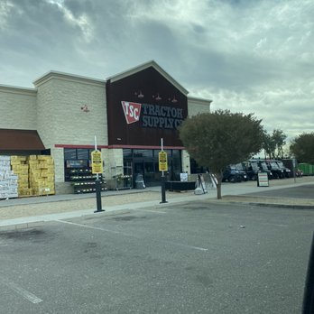 Tractor Supply - The photo captures the front facade of a Tractor Supply store, featuring a large sign with the store's name and logo. The parking lot in the foreground has marked spaces, some of which are empty. There is a small tree near the entrance, and the sky above is partly cloudy with patches of blue. The store's exterior includes a white textured wall, a black signboard, and some stacked yellow and white products near the entrance. - Department Stores Near Me - Apache Junction, Arizona