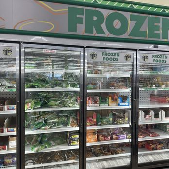 Dollar Tree - Frozen food - A grocery store freezer with three glass doors contains multiple shelves stocked with frozen food packages. The packages are in different colors and sizes, indicating a variety of frozen products. The freezer is labeled 'Frozen Foods' at the top, and a large sign above reads 'FROZEN.' The store's interior lighting reflects on the glass doors, and the background shows store aisles and lighting fixtures. - Discount Store Near Me - Catonsville, Maryland