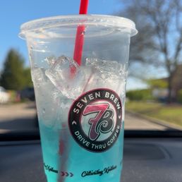 Ocean Breeze - A clear plastic cup containing a blue-colored beverage with ice cubes and a red straw is positioned on a car dashboard. The cup has a Seven Brew Coffee logo and the text 'Drive Thru Coffee' visible on it. The background shows a bright blue sky with some trees and a few clouds, indicating a sunny day.