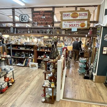 Kingston Consignments - This place is huge! - The image shows the interior of Kingston Consignments, a large consignment shop featuring multiple shelves and display units filled with antiques, ceramics, books, and collectibles. The store has a wooden floor and is well-lit, with signs hanging from the ceiling indicating parking and pricing information. Customers are seen browsing towards the back of the store, which appears organized and spacious. - Antiques Near Me - Kingston, New York