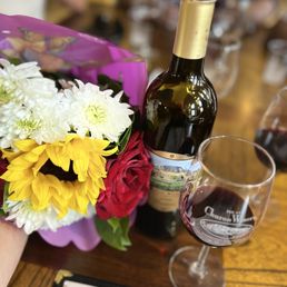 Delicious selection- Barbera Wine - A bouquet of colorful flowers, including white, yellow, and red blossoms, is positioned next to a bottle of wine with a scenic label and a glass of red wine. The setting appears to be a dining table at Churon Winery, with blurred people and glasses in the background, indicating a social gathering or celebration.