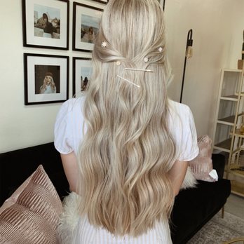 Willow Salon - The image depicts a woman with long, wavy blonde hair, styled with decorative hairpins and accessories, viewed from behind. She is wearing a light-colored, striped dress or top. The background features a wall with multiple framed photographs, a black sofa with pink and white pillows, and a modern floor lamp, indicating an indoor setting, possibly a salon or a home interior. - Hair Salons Near Me - Tucson, Arizona