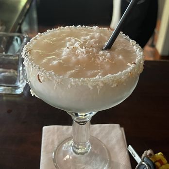 El Bruno's - Coconut Marg! Loved it and my El Bruno beer I had waiting for my to go order - A creamy cocktail with a salted rim and a black straw is presented in a large, elegant glass goblet. The drink appears smooth and frothy, sitting on a white napkin on a dark wooden surface. The background suggests a bar or restaurant setting with blurred elements and a person partially visible. - Mexican Near Me - Albuquerque, New Mexico