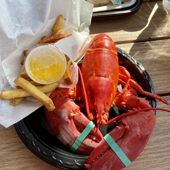 Champlin's Seafood - 1 1/2 lb lobster dinner - A seafood meal includes a large cooked lobster on a black tray, accompanied by French fries with a small cup of melted butter. The meal is served on a wooden table outdoors, with drinks visible in the background, including a glass of beer and a glass of water. The setup suggests a casual dining experience, likely at a seafood restaurant or outdoor eatery. - Seafood Near Me - Narragansett, Rhode Island