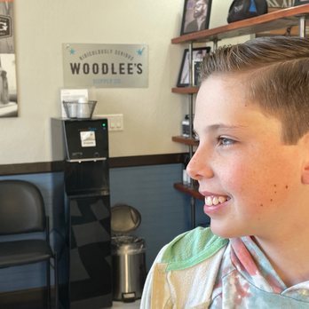Precision Cut - A boy with short, neatly styled hair and freckles is smiling and looking to the right in an indoor setting. The background includes shelves with framed photos, a water dispenser, and a wall sign that reads 'Woodlee's Supply Co.' The environment appears to be a salon or barbershop, with a chair and other salon-related items visible. - Hair Salons Near Me - Gilbert, Arizona