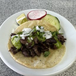 carne asada taco - A carne asada taco on a corn tortilla is topped with chopped onions and cilantro. The plate includes slices of cucumber, radish, and a lime wedge, all placed on a gray surface. The presentation highlights traditional Mexican flavors and fresh ingredients.