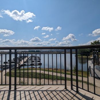Catfish Charlie's - a view of a dock from a balcony - The image shows a river viewed from a balcony with a metal railing. There is a dock with multiple boat slips extending into the river, and several boats are moored along the shoreline. A walkway runs parallel to the river, and the sky is partly cloudy with scattered white clouds. The scene includes some greenery and distant structures, indicating a calm, outdoor setting near a marina or waterfront area. - Seafood Near Me - Dubuque, Iowa