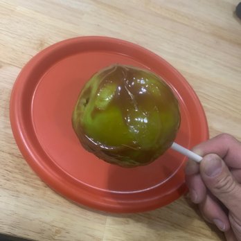 The Snack Shack - a person dipping a chocolate covered apple on a red plate - A caramel apple covered with green caramel is held by a hand on a stick. The apple is placed on a red plastic tray, which rests on a light-colored wooden table. The caramel coating appears glossy and smooth, with some uneven areas. The background shows part of the table and a dark object in the upper right corner. - Candy Stores Near Me - Keystone, Colorado