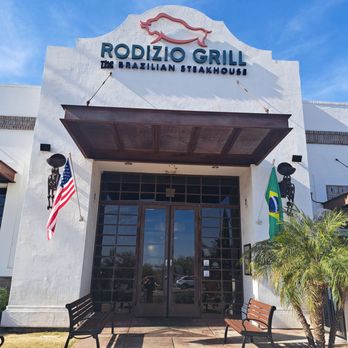 Rodizio Grill - Mesa - Exterior - The image shows the front entrance of Rodizio Grill, a Brazilian steakhouse, with flags, benches, and a white building facade. - Steakhouses Near Me - Mesa, Arizona