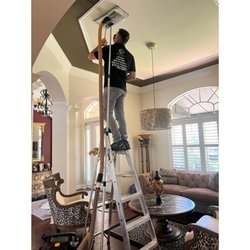 Clean Air Pros - HVAC professional on a ladder cleaning an overhead air vent in a residential living space as part of an air duct cleaning service.