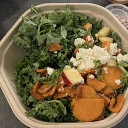 A mixed salad in a takeout container includes kale, roasted sweet potatoes, apple slices, garlic cloves, and crumbled cheese.