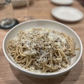The Deliciouser - Cacio e Pepe. Very tasty but a bit heavy on the Pepe. And I do love well spiced and seasoned food. Huge portions though! - A white bowl filled with cooked spaghetti topped with grated cheese is positioned on a wooden table. In the background, there are two empty white plates, a glass with a drink, and a blurred setting, suggesting a dining environment. The image focuses on the pasta dish, highlighting the cheese topping and the table setting. - Herbs & Spices Near Me - Madison, Wisconsin